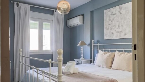 A bedroom with a white metal bed frame, a bedside table with a lamp, and a window with white curtains. - Live the charms of Seville in Triana (Sevilla)