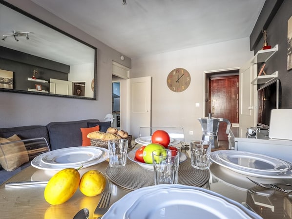 A modern living room with a dining area, featuring a sofa, a coffee table with fruit and glasses, and a kitchen visible in the background.