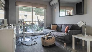 A modern living room with a grey sofa, a small white table, and a hanging chair. A large window provides a view of buildings outside. - Live the charms of Seville in Triana (Sevilla)