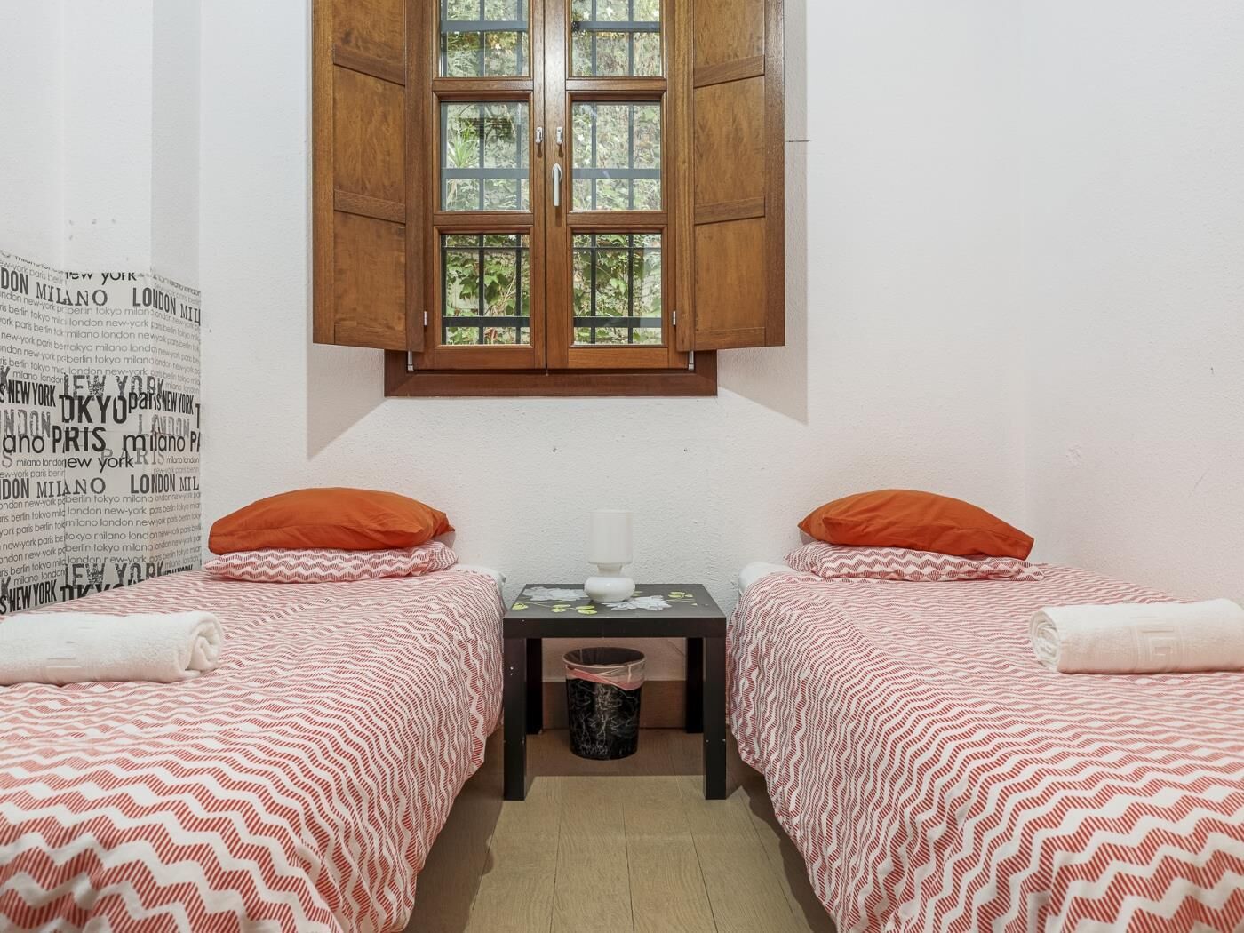 A small room with two beds, each with an orange pillow, a small table with a lamp, and a window with wooden shutters.