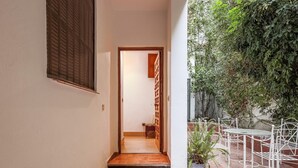 Un couloir étroit avec une porte en bois qui mène à une pièce. On aperçoit une plante en pot sur le côté droit et une chaise en métal au premier plan.