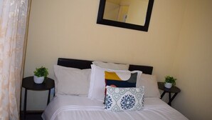 Una cama bien hecha con ropa blanca, un cabecero negro y varios almohadones. Hay dos mesitas de noche con plantas en macetas. Un espejo cuelga sobre la cama.