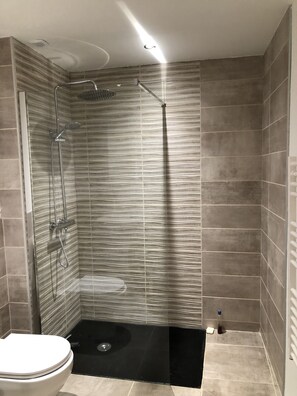 Bathroom features a shower with a glass door and handheld showerhead, surrounded by large tiles and a dark floor.
