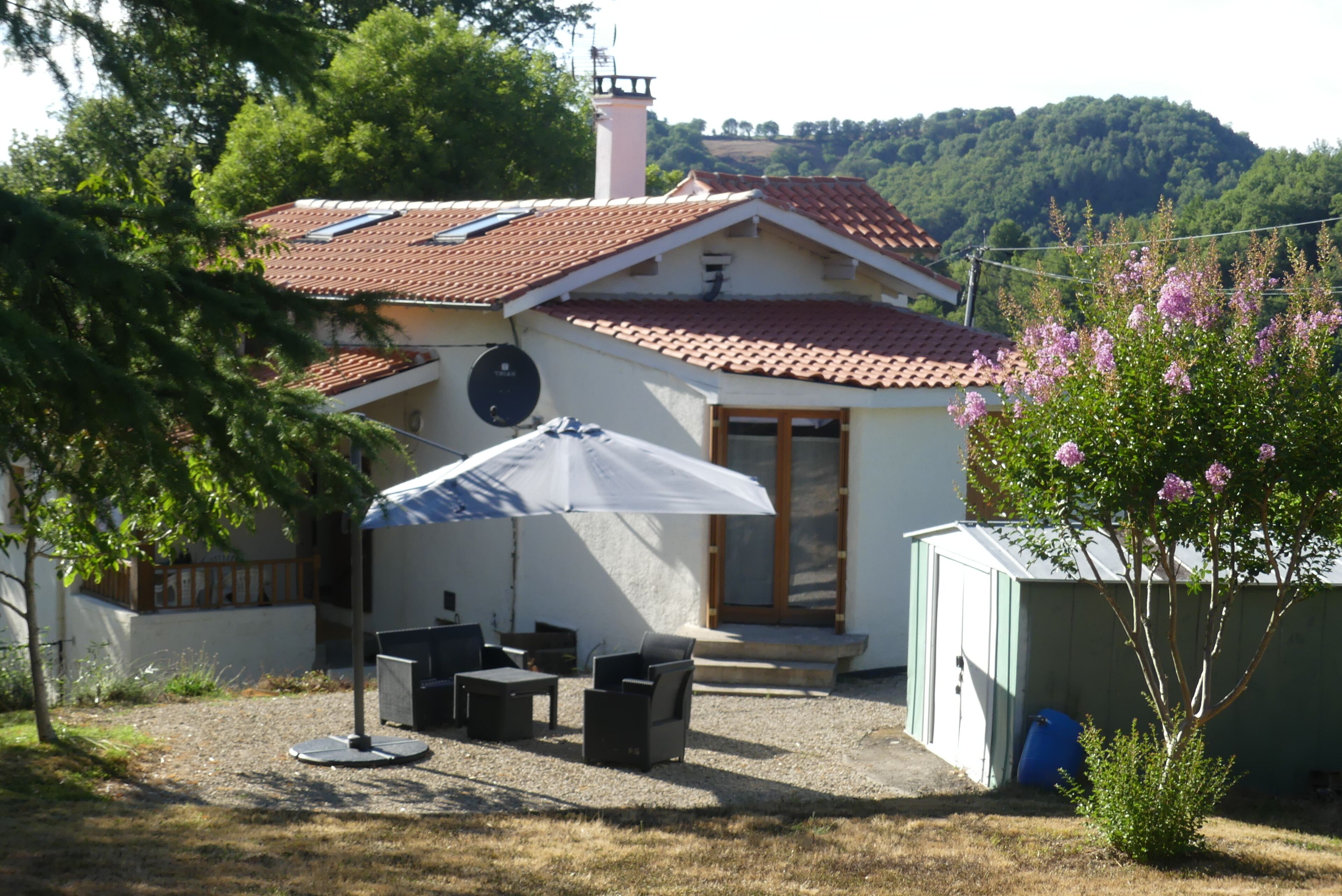 Une maison avec un toit en tuiles, un patio couvert avec des meubles d’extérieur et un petit jardin agrémenté de plantes à fleurs.