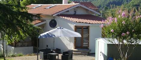 Una casa con techo de tejas, un porche cubierto con mobiliario exterior y un pequeño jardín con plantas en flor.