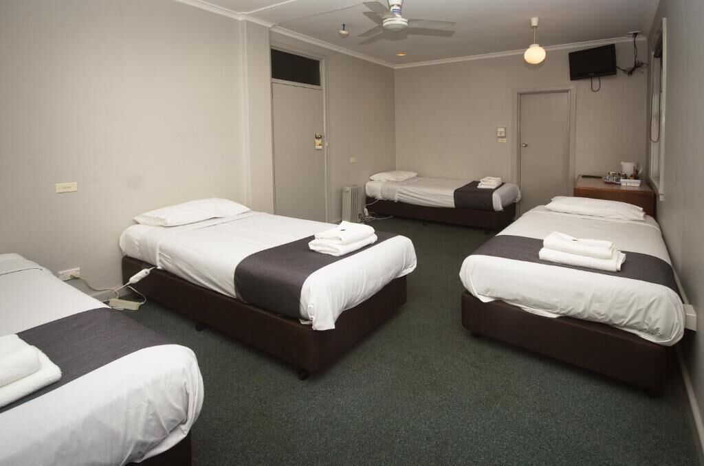 Habitación de hotel con tres camas, ventilador de techo, televisión y una mesita de noche con lámpara.