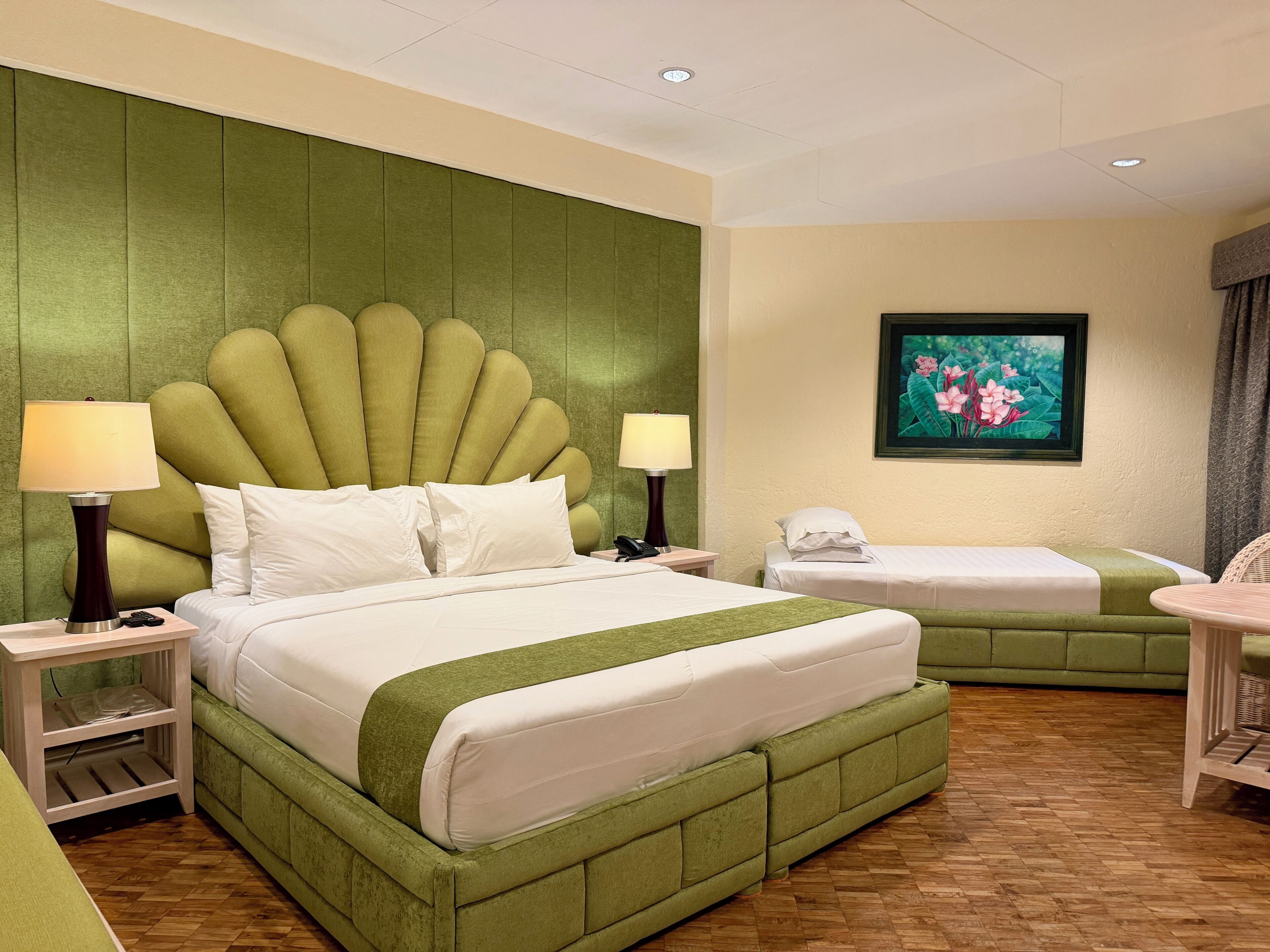 A hotel room with a large bed, two bedside tables, a small table, a green upholstered headboard, a framed picture on the wall, and a wooden floor.