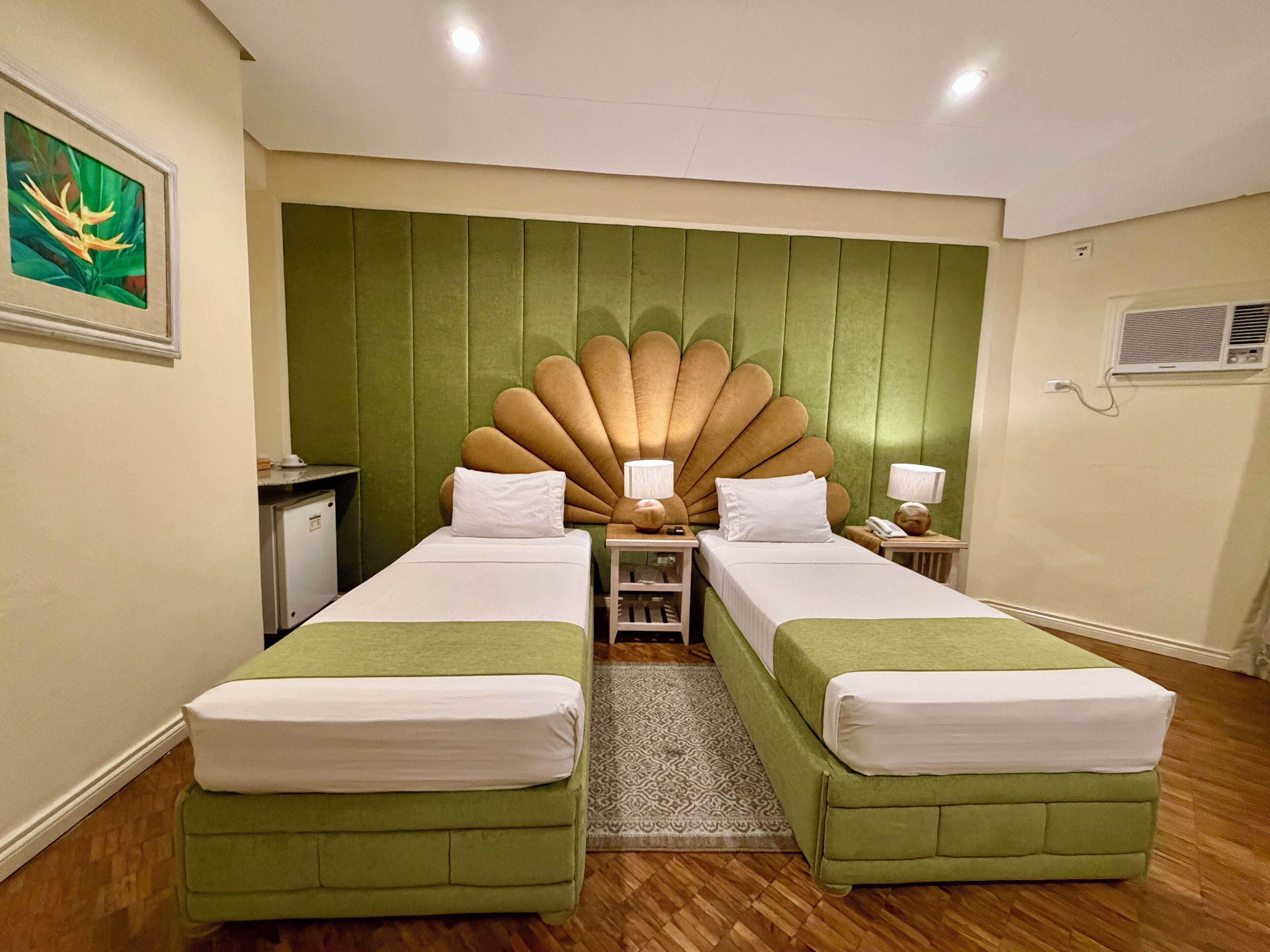 A hotel room with two single beds, a green upholstered headboard, bedside tables with lamps, a small desk, and a framed picture on the wall.