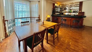 A dining area with a wooden table and chairs, a bar with shelves of bottles, and a view through large windows with curtains. - Coco Grande Hotel (Dumaguete)