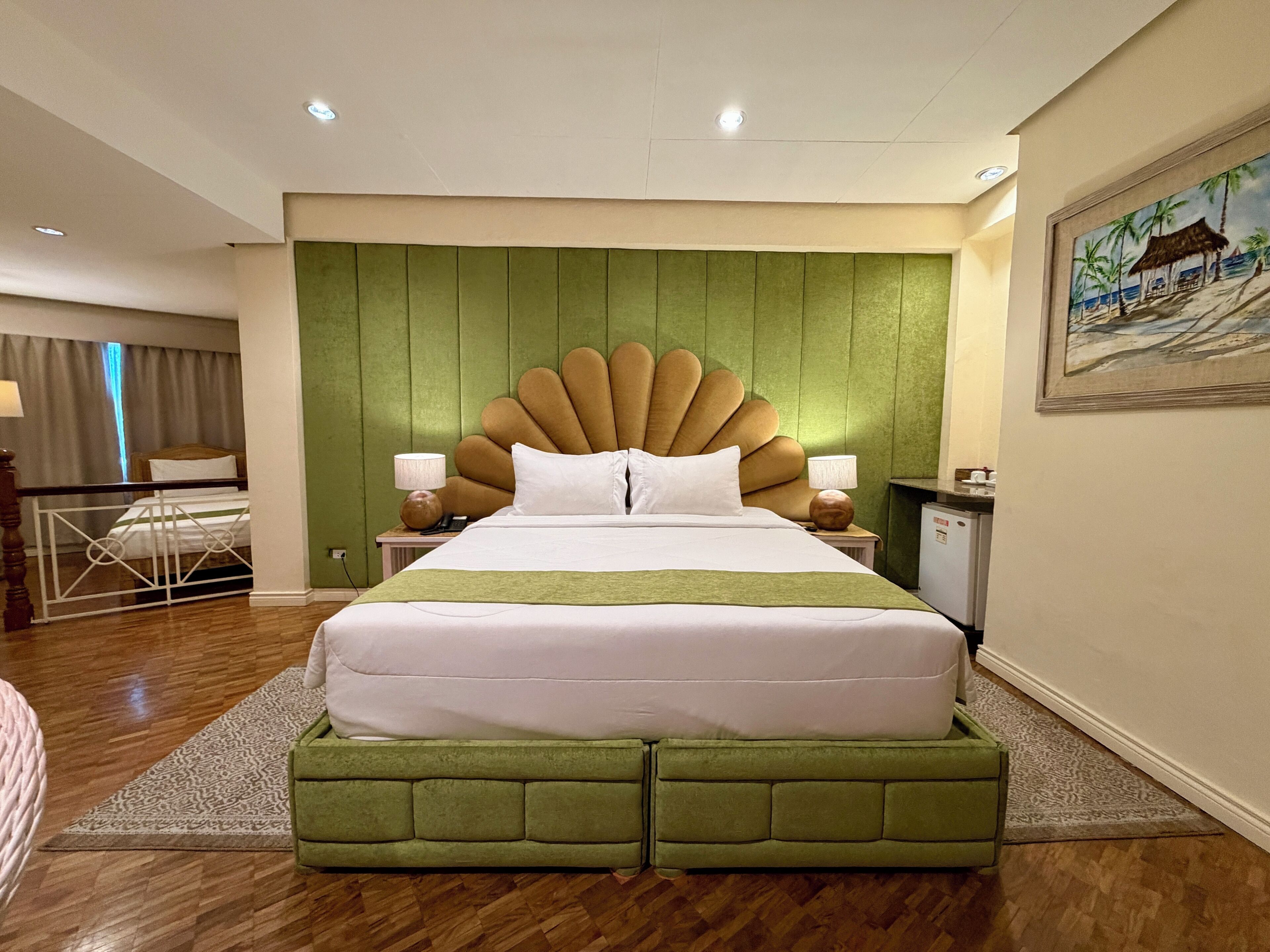 A hotel room with a large bed, green headboard, two bedside tables with lamps, a small desk, and a painting on the wall.