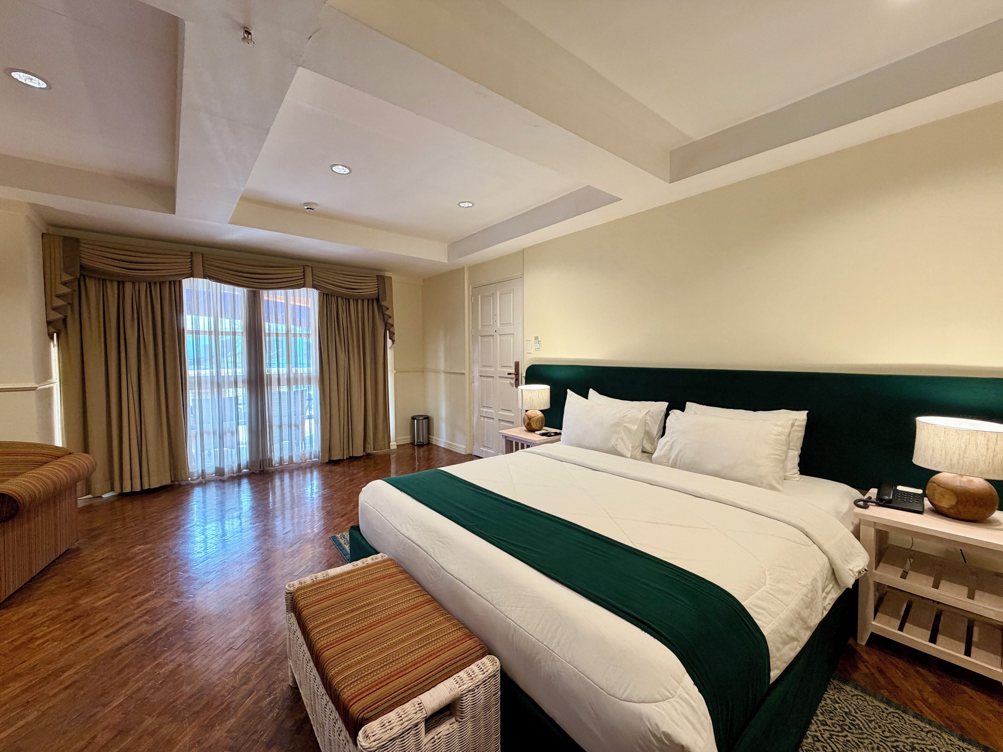 A hotel room with a large bed, a nightstand, a wicker chair, and a view of the outdoors through the window.