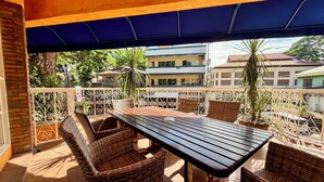 A balcony with a wooden table and wicker chairs, a blue awning, and a view of buildings and trees. - Coco Grande Hotel (Dumaguete)