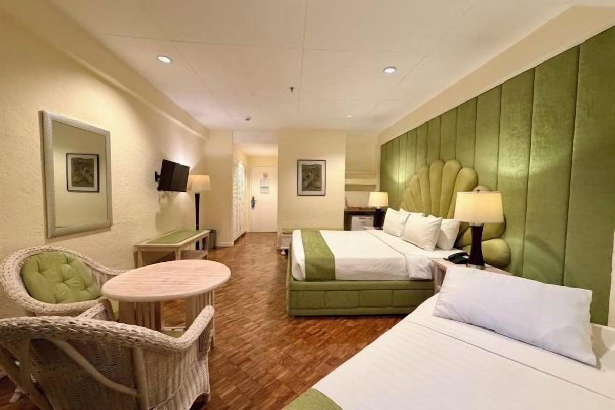 A hotel room with a green upholstered headboard, a single bed, a small round table, wicker chairs, and a television.
