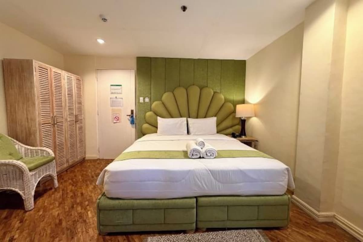 A hotel room with a large bed, a green upholstered headboard, a white chair, and a wardrobe.