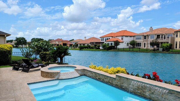 A swimming pool with a stone edge, surrounded by a patio with lounge chairs. In the background, there are houses with red roofs and a body of water. - Villa Oak Lake Azure - Ultra Luxury Lakeview Oasis (Houston)