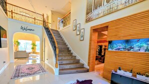 A spacious foyer with a staircase, a welcome sign, a large aquarium, and a cowhide rug. - Villa Oak Lake Azure - Ultra Luxury Lakeview Oasis (Houston)