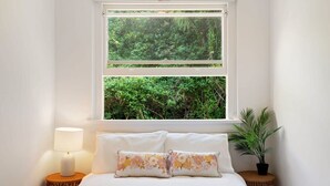 Une chambre à coucher avec un lit, deux oreillers, une lampe de chevet, une fenêtre donnant sur un paysage verdoyant et une petite plante en pot.