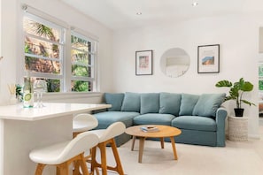 Un salon moderne avec un canapé bleu clair, une table basse blanche et un coin bar blanc avec deux chaises blanches. Deux tableaux encadrés ornent le mur, et une plante en pot se trouve sur la droite.