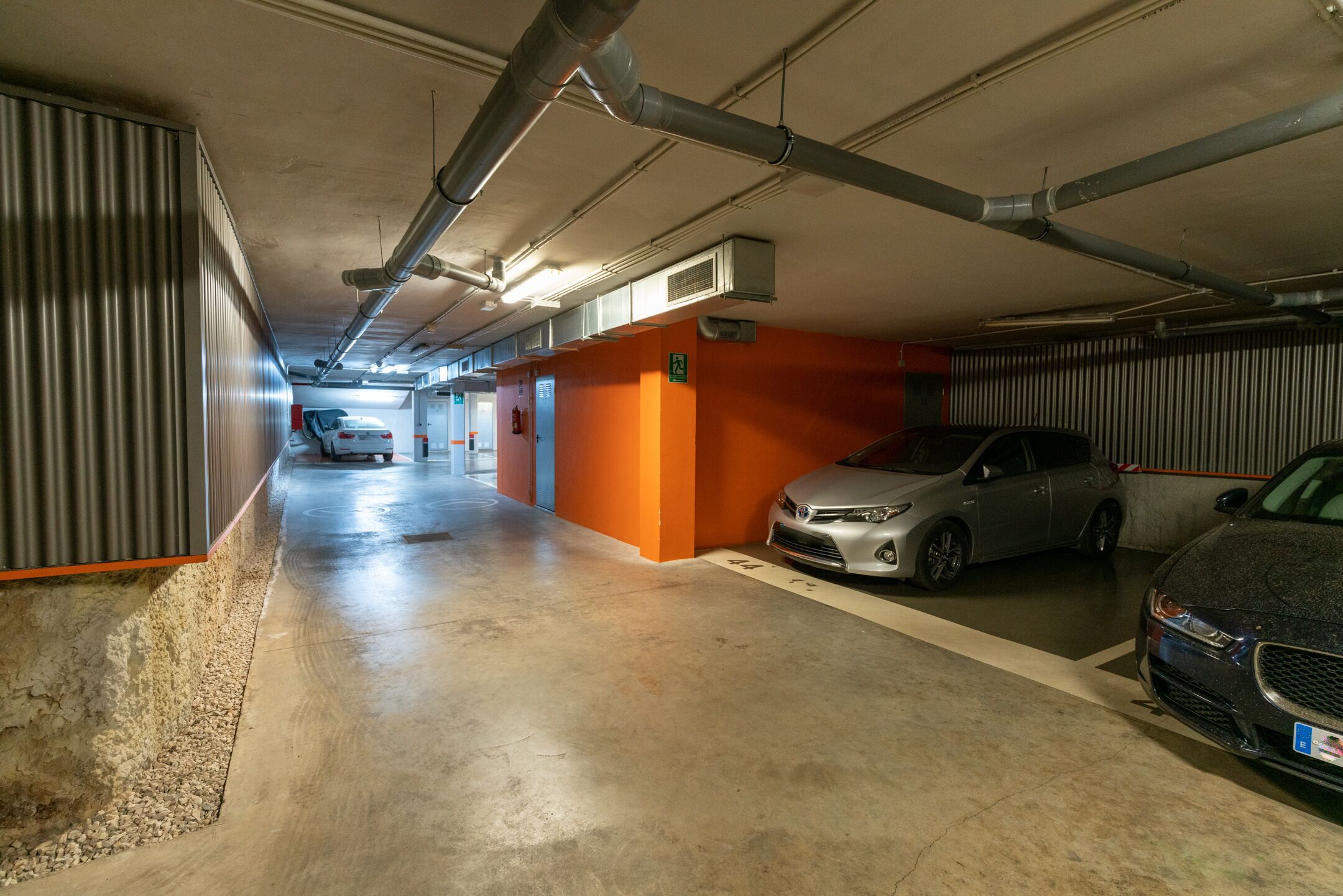 Un estacionamiento con coches aparcados en espacios designados, suelo de hormigón y conductos de ventilación visibles.