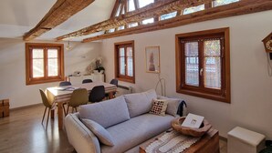 Uma sala de estar ampla com teto alto, vigas de madeira, ventilador de teto e grandes janelas que permitem a entrada de luz natural.