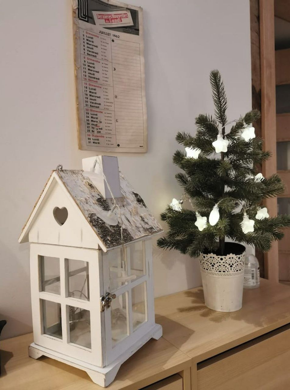 Un pequeño comedero para pájaros blanco con ventana en forma de corazón y tejado, sobre una superficie de madera junto a un árbol de Navidad iluminado.