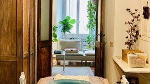 Uma sala de jantar com mesa de madeira, cadeiras brancas, um centro de mesa com uma tigela verde, e vista para uma sala de estar com sofá e plantas.