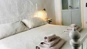 Cama bem arrumada com travesseiros, abajur e toalhas dobradas em cabeceira de metal branca.