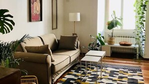 Uma sala de estar com sofá, mesa de centro, tapete estampado e janela com plantas.