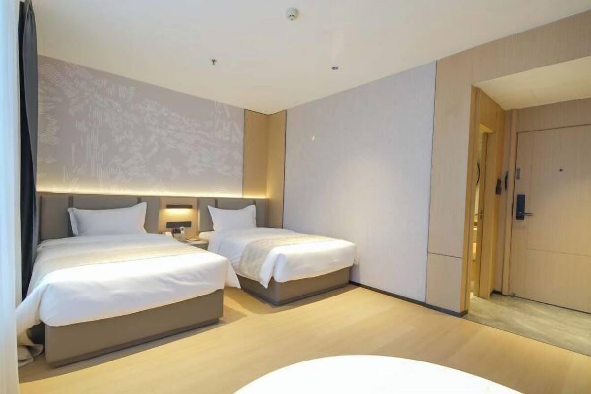 A hotel room with two beds, a nightstand, a wall-mounted lamp, and a door to the right.