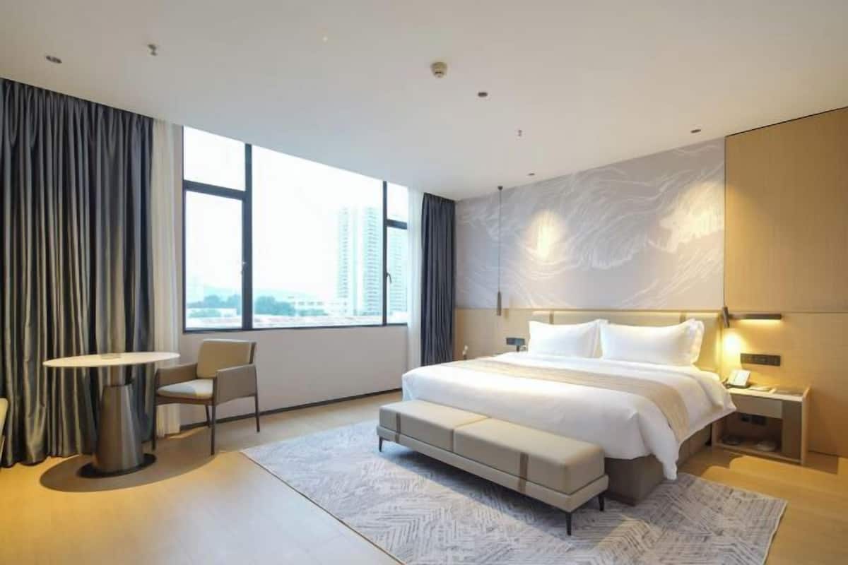A modern hotel room with a large bed, a desk, a chair, and a view of the city.