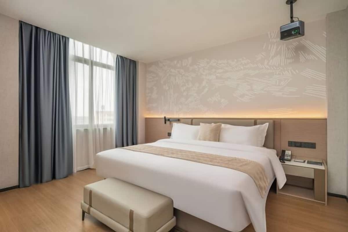 A hotel room with a large bed, a desk, a projector, and a window with curtains.