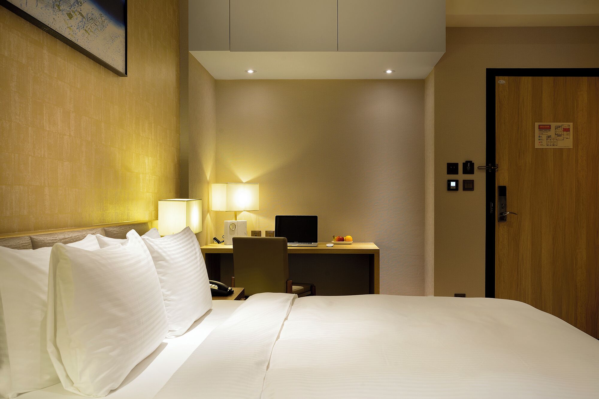 A hotel room with a bed, a desk with a laptop, a lamp, and a door with a lock and keycard reader.