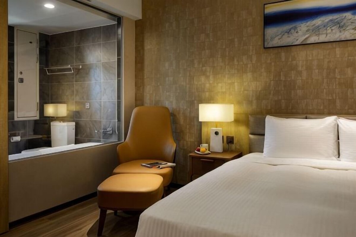 Une chambre d’hôtel comprenant un lit, une chaise, une table de chevet, une lampe, une salle de bain avec douche et une grande fenêtre.