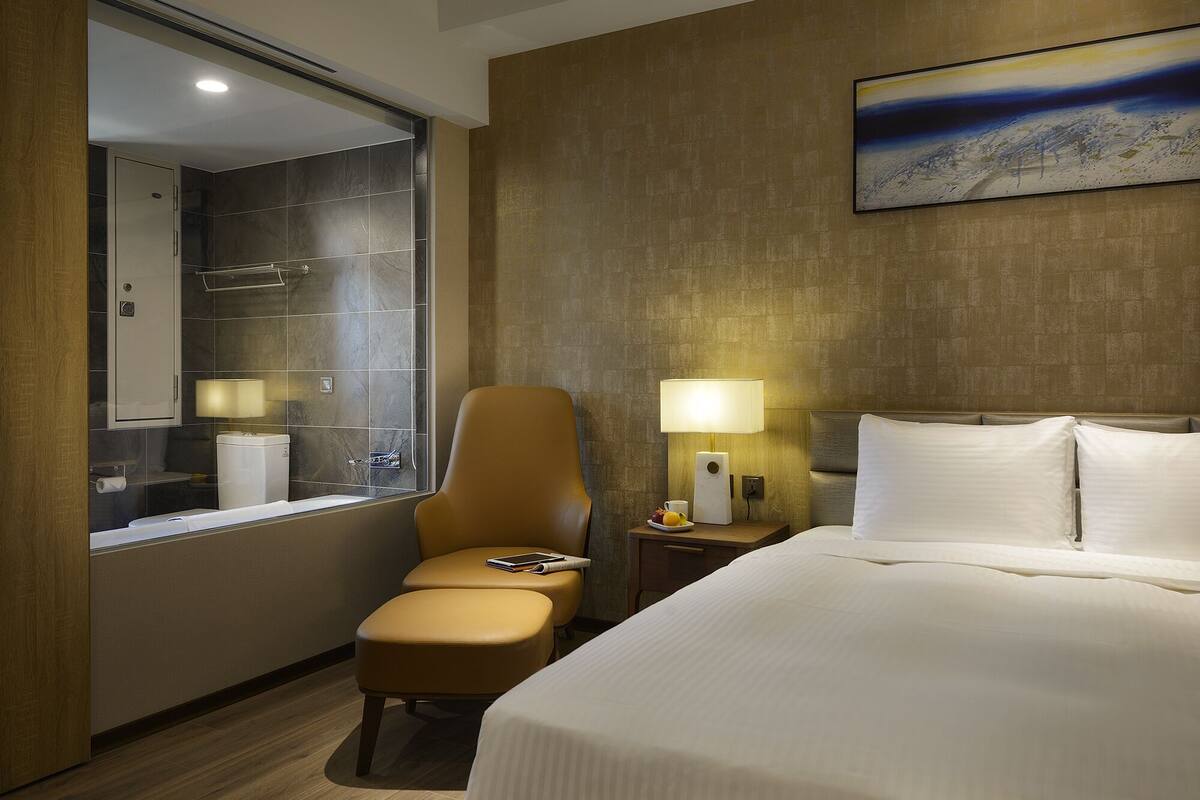 Habitación de hotel con cama, una silla, mesita de noche con lámpara, baño con ducha y un cuadro en la pared.