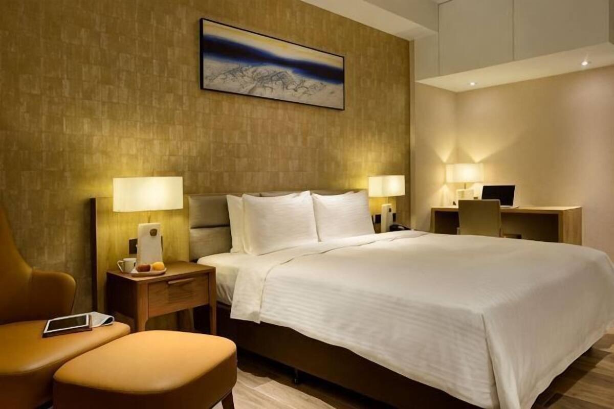 Una habitación de hotel con una cama, dos mesitas de noche con lámparas, una silla, un escritorio y un cuadro en la pared.
