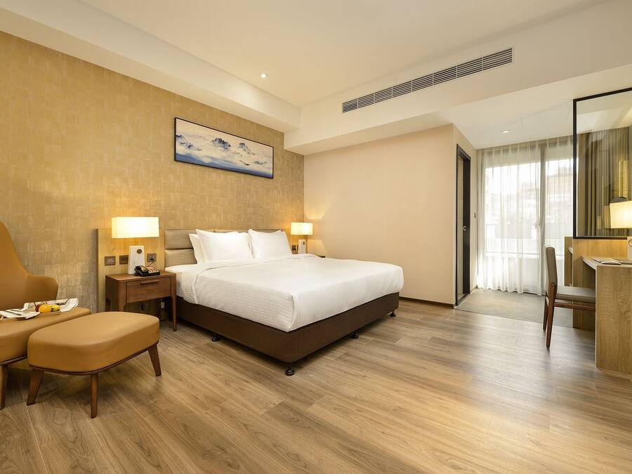 A hotel room with a large bed, two bedside tables with lamps, a chair, a desk with a lamp, and a large window with curtains.
