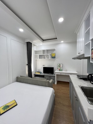 A modern, compact hotel room with a bed, a small kitchenette, a TV, and a desk area.