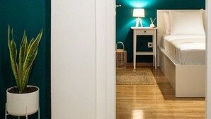 Una habitación con paredes color turquesa, una cama blanca, una mesita de noche con una lámpara y una planta en maceta.