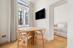 A modern room with a round wooden table, two chairs, a flat-screen TV mounted on the wall, and a bed with white bedding.