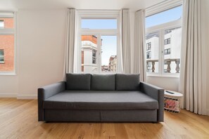 A grey sofa in a room with large windows, wooden flooring, and a small table with a remote control.