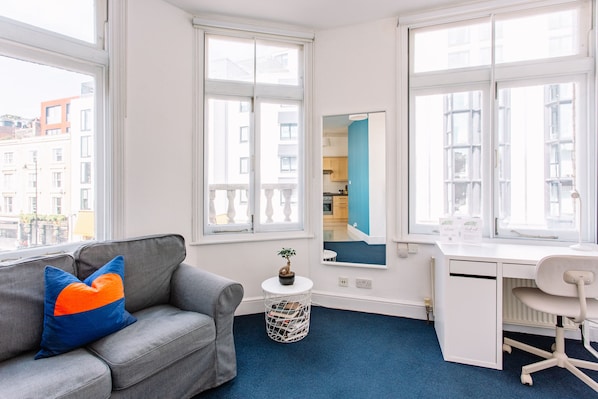 A modern room with a grey sofa, a small round table with a plant, a white desk with a chair, and large windows offering a city view.
