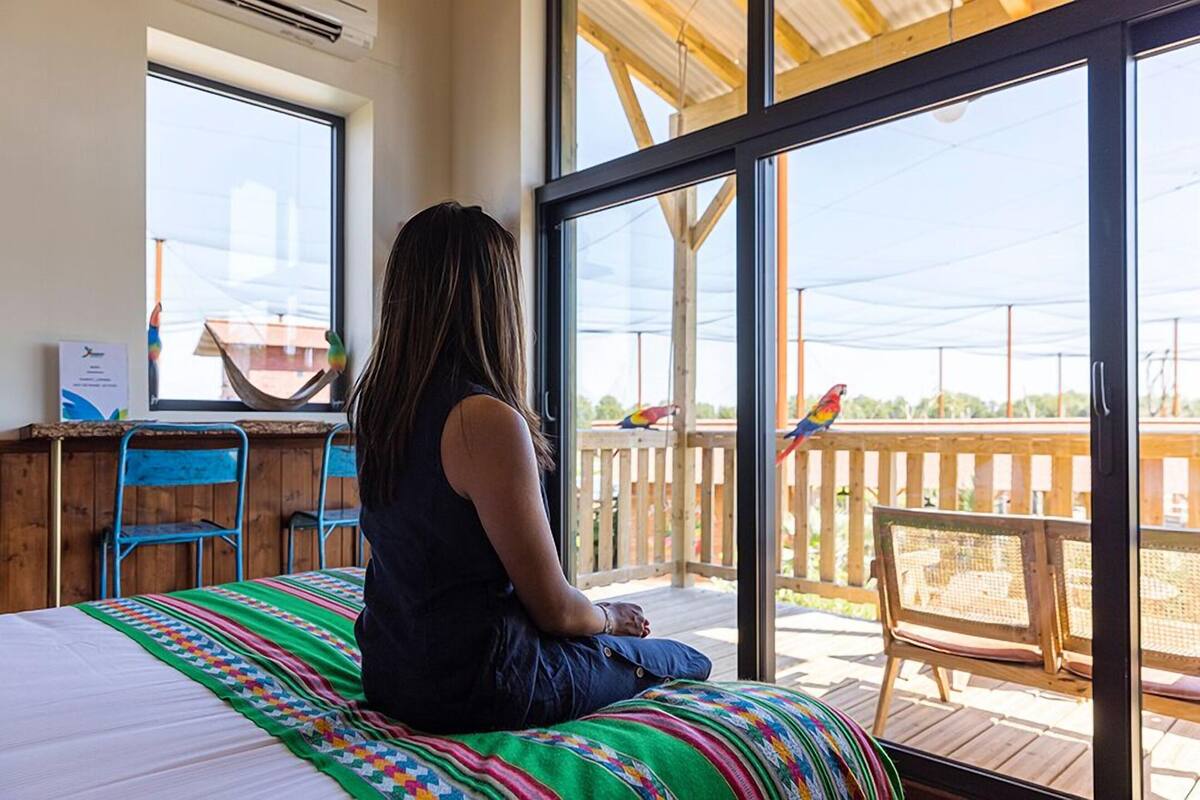 A person sitting on a bed with a colorful blanket, looking out of a large sliding glass door at a wooden deck with chairs and a bird.