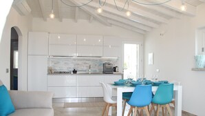 A modern kitchen with white cabinets, a tiled backsplash, and a dining area with a white table and blue chairs. - R House (Budoni)