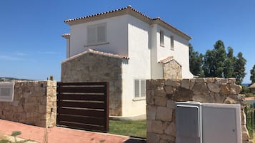 Un edificio blanco con base de piedra y techo de tejas, que cuenta con una entrada con reja y una pequeña ventana.