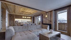 A modern living room with a large sectional sofa, a wooden coffee table, and a view of a kitchen and mountains through large windows. - Spacious apartment near slopes with jacuzzi and parking (Montvalezan-La Rosière)