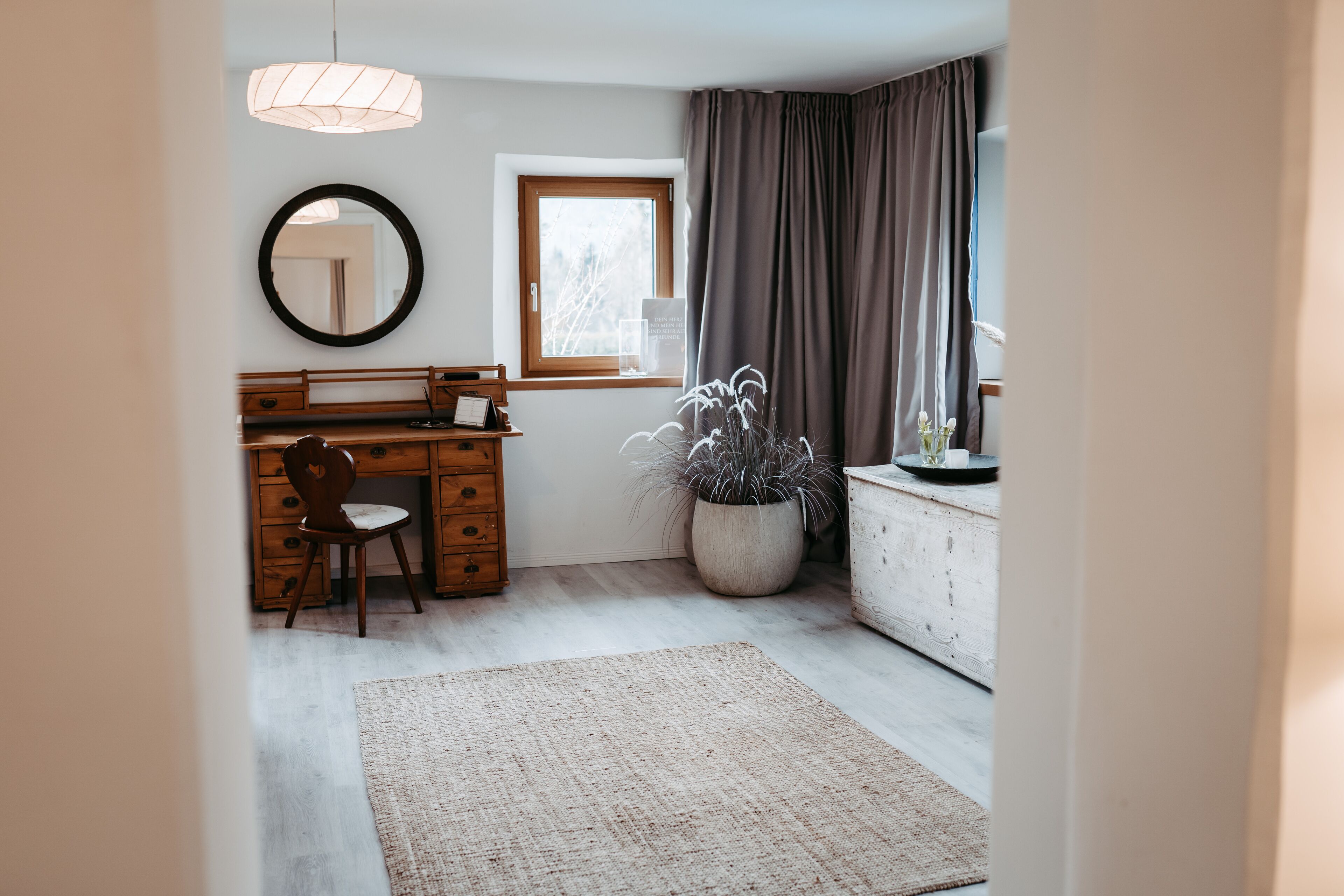 Une chambre avec une commode en bois, un miroir rond, une grande plante en pot, un bureau avec une chaise et une fenêtre avec des rideaux.