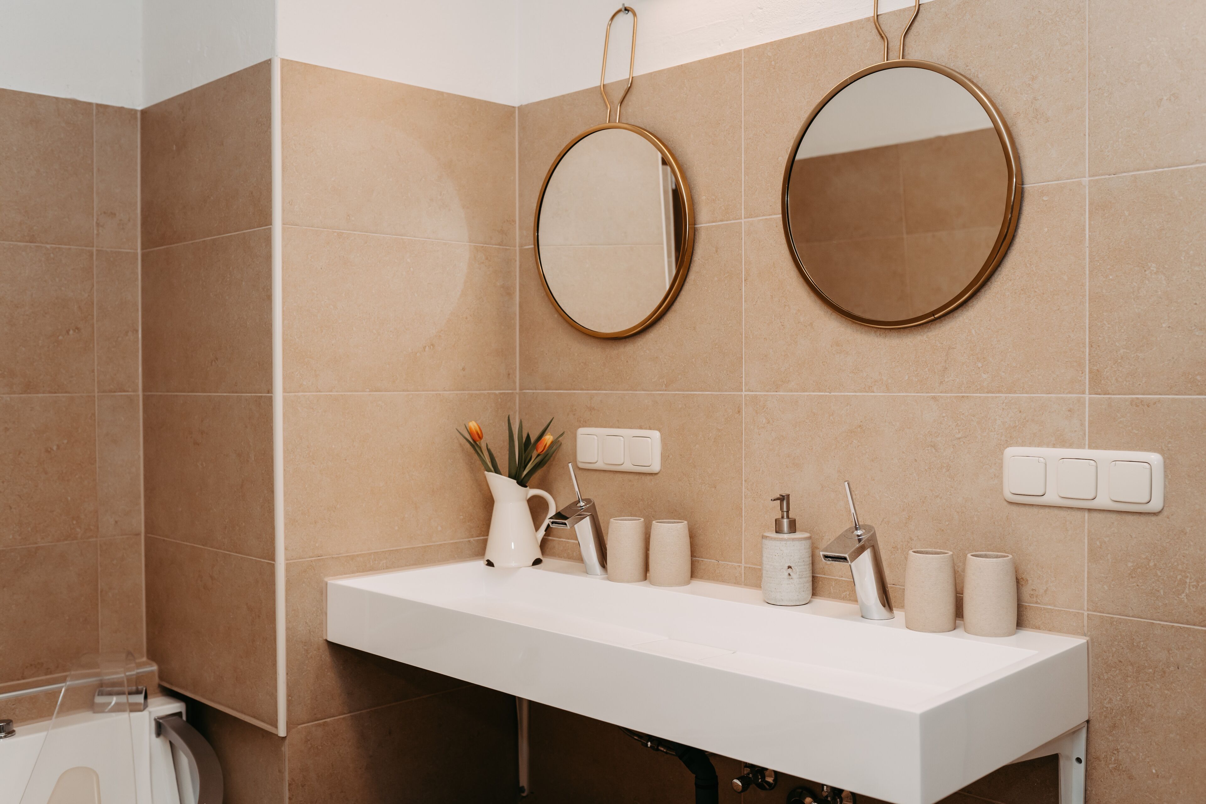 A bathroom with a white sink, two round mirrors, a white vase with flowers, and a towel rack.