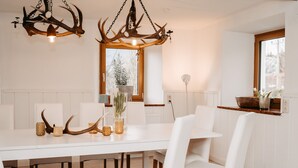 Een eetruimte met een witte tafel, witte stoelen, een lamp met gewei en een raam met uitzicht op bomen.