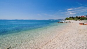 Una spiaggia limpida con acque calme, riva di ciottoli e alcuni edifici sullo sfondo.