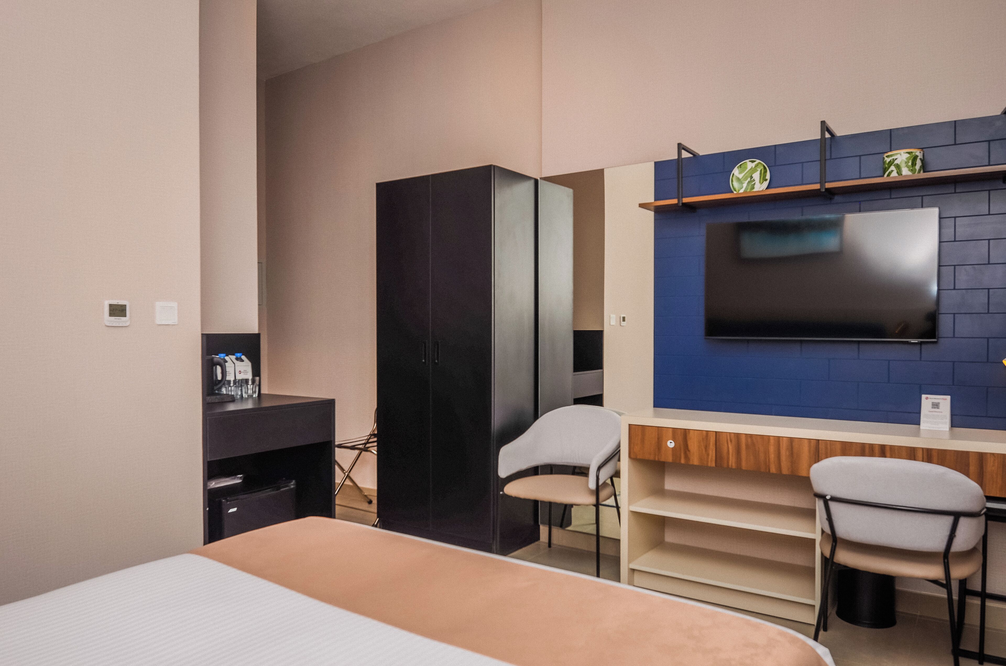 Una habitación de hotel moderna con una cama, un escritorio con dos sillas, un armario, un televisor de pantalla plana montado en una pared de ladrillo azul y un estante pequeño sobre el televisor.